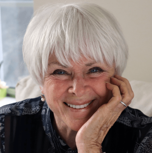 Byron Katie with short white hair is smiling, resting one hand on her cheek, while wearing a patterned top and pearl earrings.